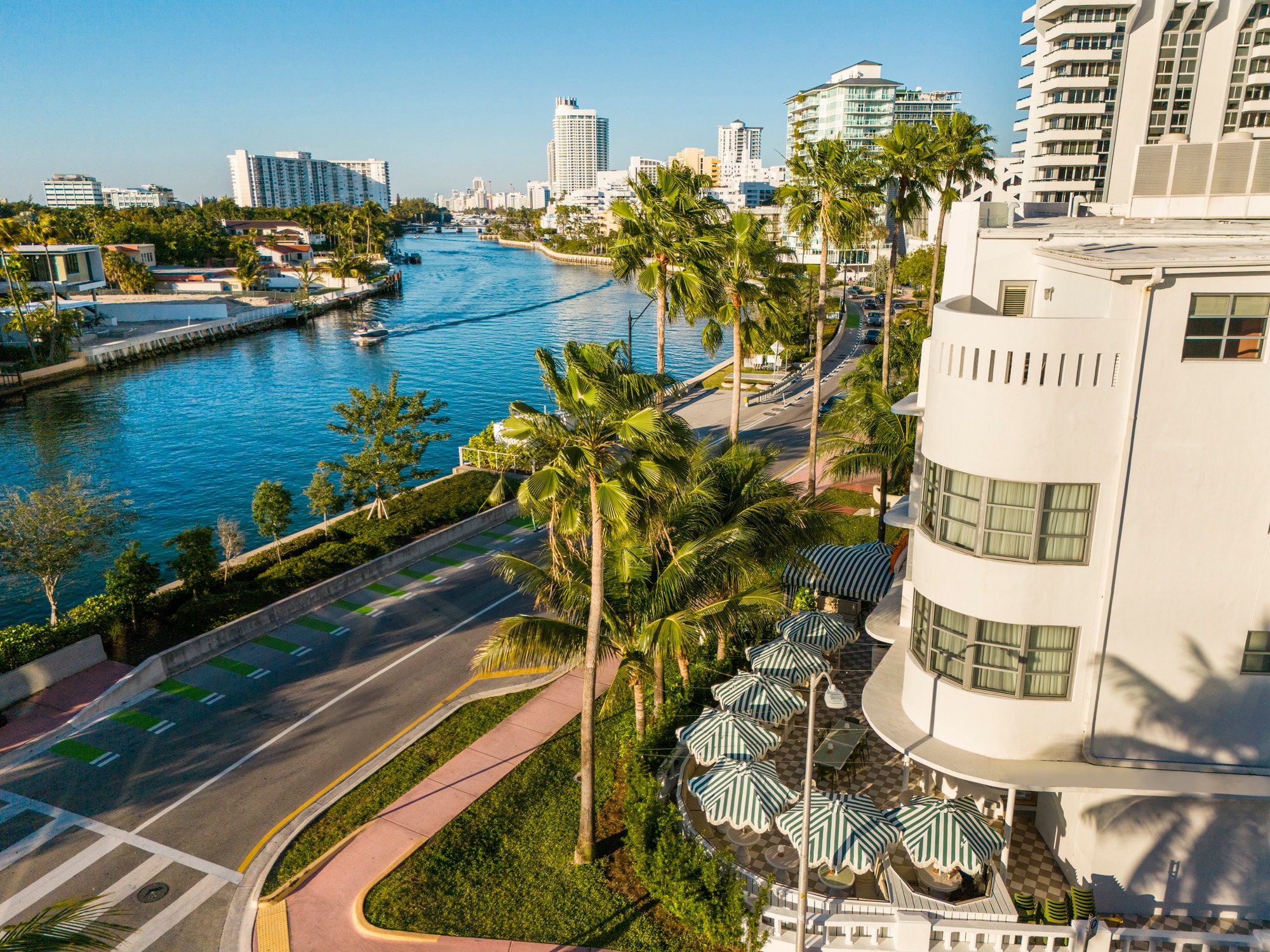 Aerial view of a white building with palm trees and umbrellas in front of it, with a road and waterway in the background.
