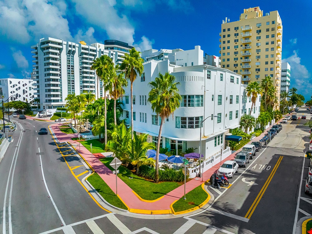 A white, multi-story building with palm trees in front, on a street corner with parked cars and a blue sky.