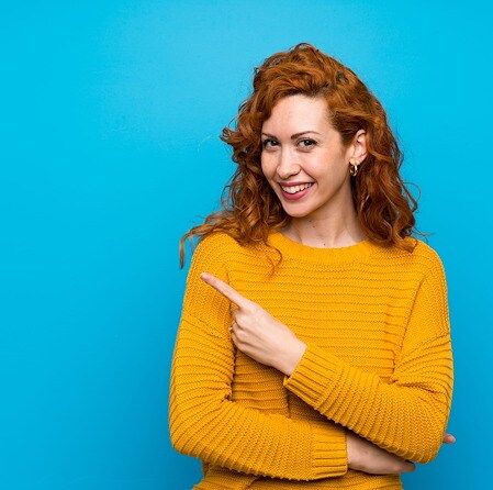 Woman with curly red hair wearing a yellow sweater pointing to the side against a blue background, representing career opportunities at Air Academy Credit Union