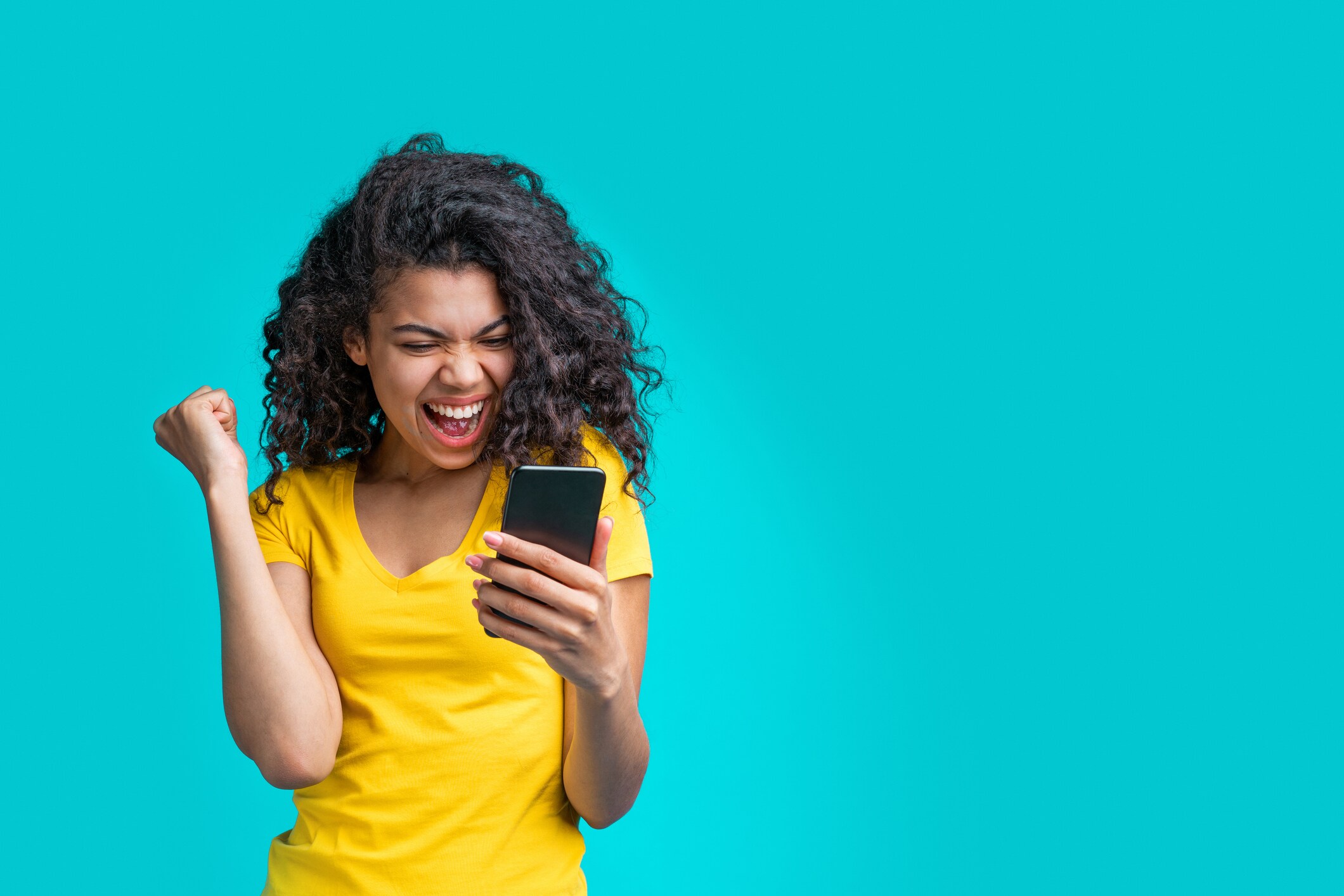 Joyful young woman in a yellow shirt cheers with a raised fist while reading her smartphone, smiling against a bright turquoise background.