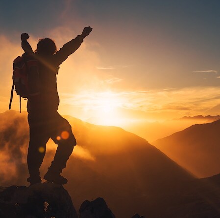 A silhouette of a hiker with a backpack standing on a mountain peak with arms raised against a bright, golden sunset sky and misty mountain ranges.