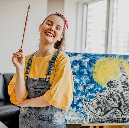 Smiling female artist in a yellow shirt and denim overalls holds a paintbrush beside a vibrant blue-and-yellow painting in a bright studio.
