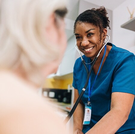Smiling Black healthcare worker in blue scrubs and a stethoscope leans toward a blurred patient, creating a warm, reassuring clinical visit.