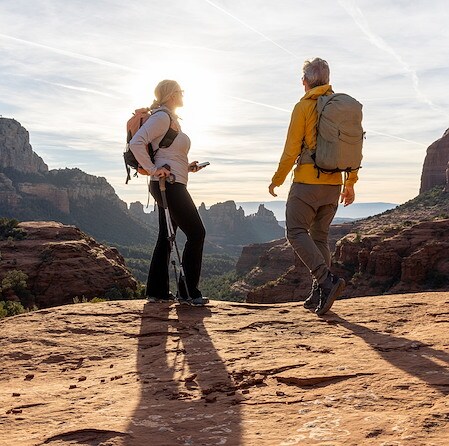 A couple with backpacks stands on a red rock cliff overlooking a desert mountain landscape during a bright, sunny day with long shadows cast behind.