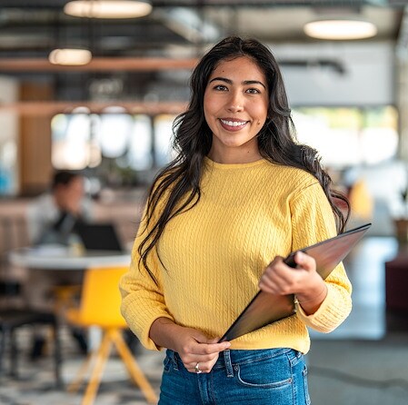 Smiling professional woman in a yellow sweater holds a tablet in a bright, modern coworking office, conveying confidence and warmth.