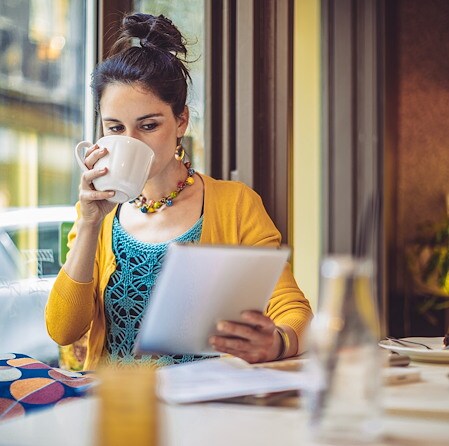 Woman sitting near a window in a cafe drinking from a mug while looking at a tablet