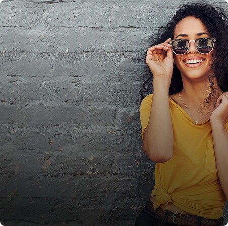 Smiling woman wearing sunglasses and a yellow top standing against a grey brick wall