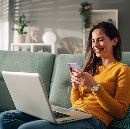 A woman in a yellow sweater smiles while using a smartphone, sitting on a green couch with a laptop open in front of her.