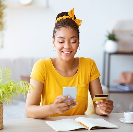 Smiling Young Woman Making Online Purchase With Card And Phone At Desk