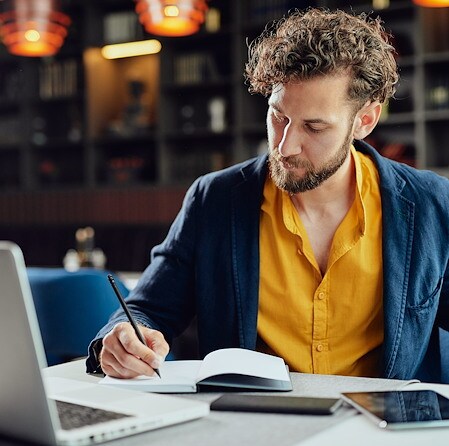 A man with curly hair and a beard writing in a notepad sitting in front of an open laptop