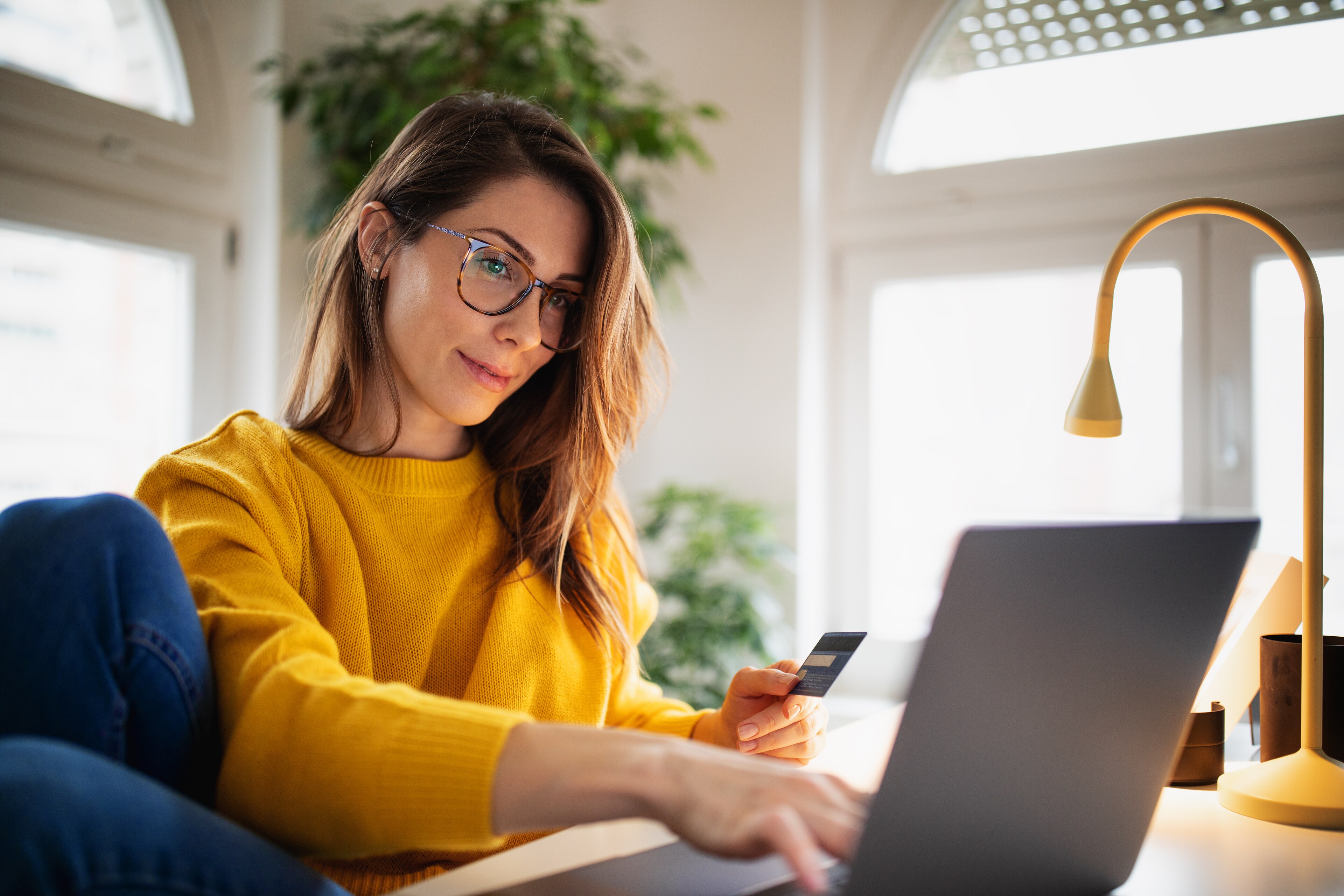 Woman with glasses in a yellow sweater holding a debit card while typing on her laptop at home