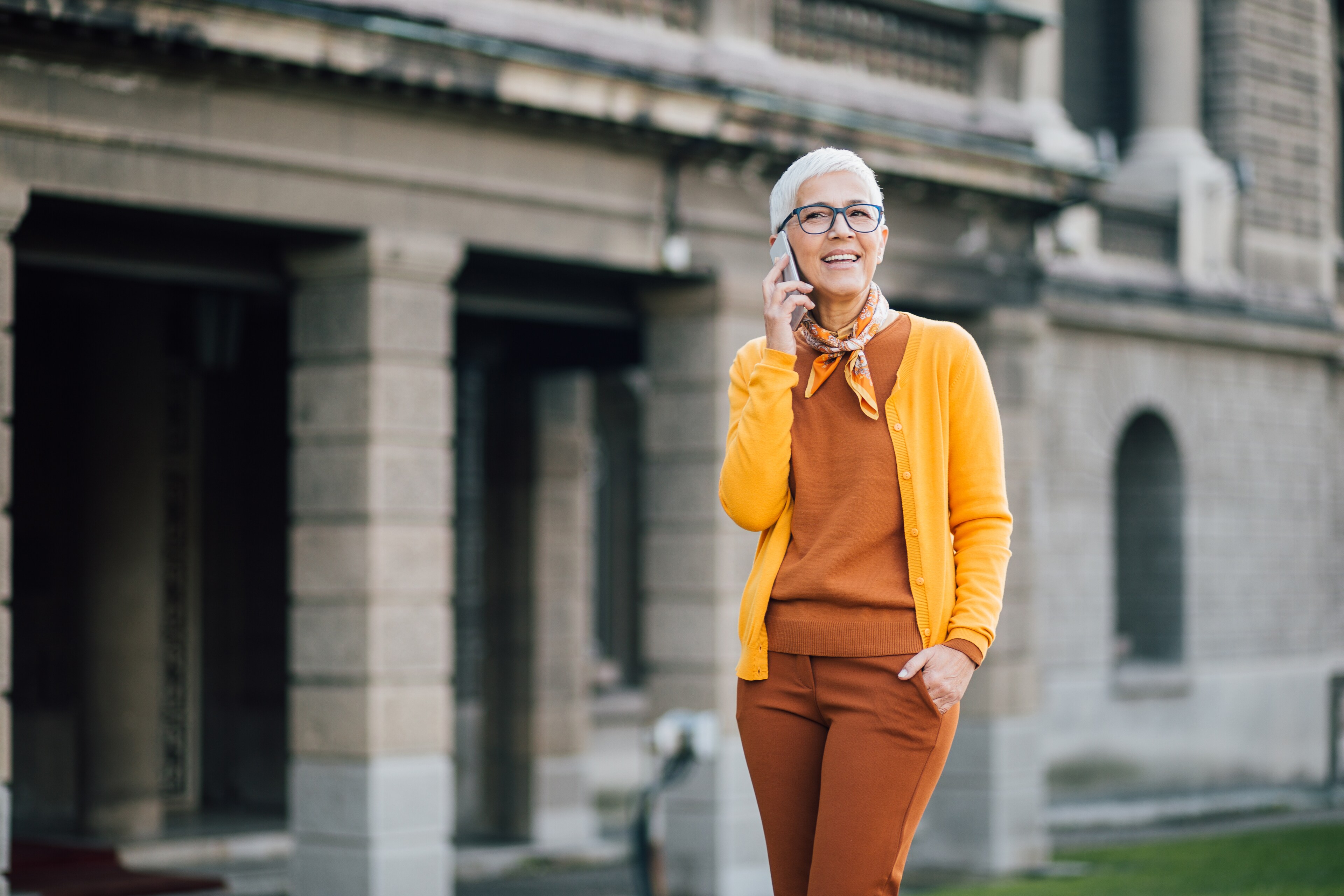 Stylish senior woman with short white hair wearing glasses and a yellow cardigan talking on a mobile phone outside a historic stone building