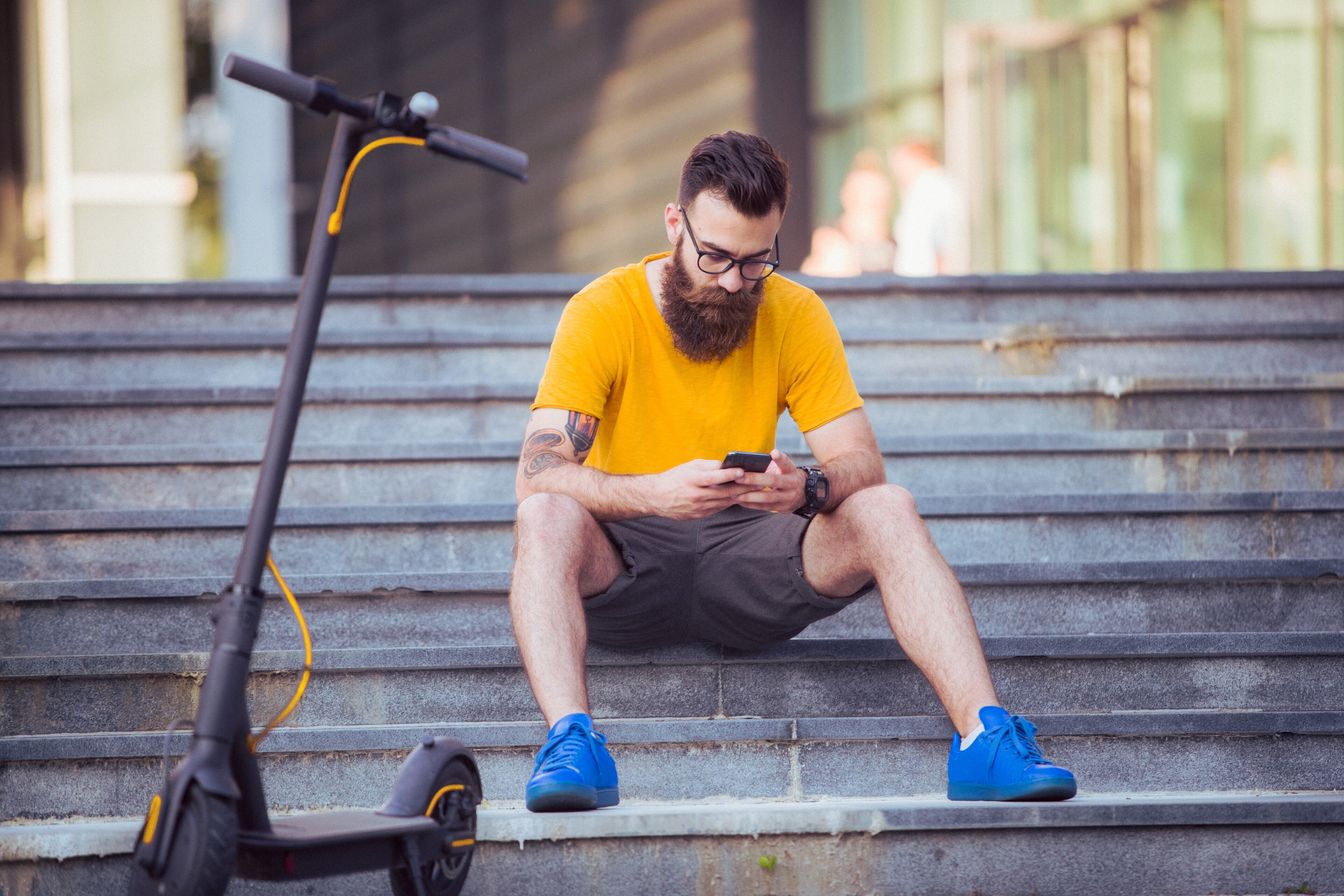 Bearded man in a yellow t-shirt using his phone while sitting on outdoor steps near an electric scooter