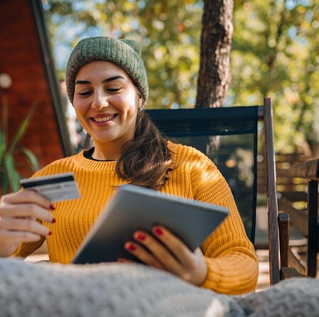 Smiling woman wearing a green beanie and mustard sweater sitting outdoors while holding a credit card and tablet