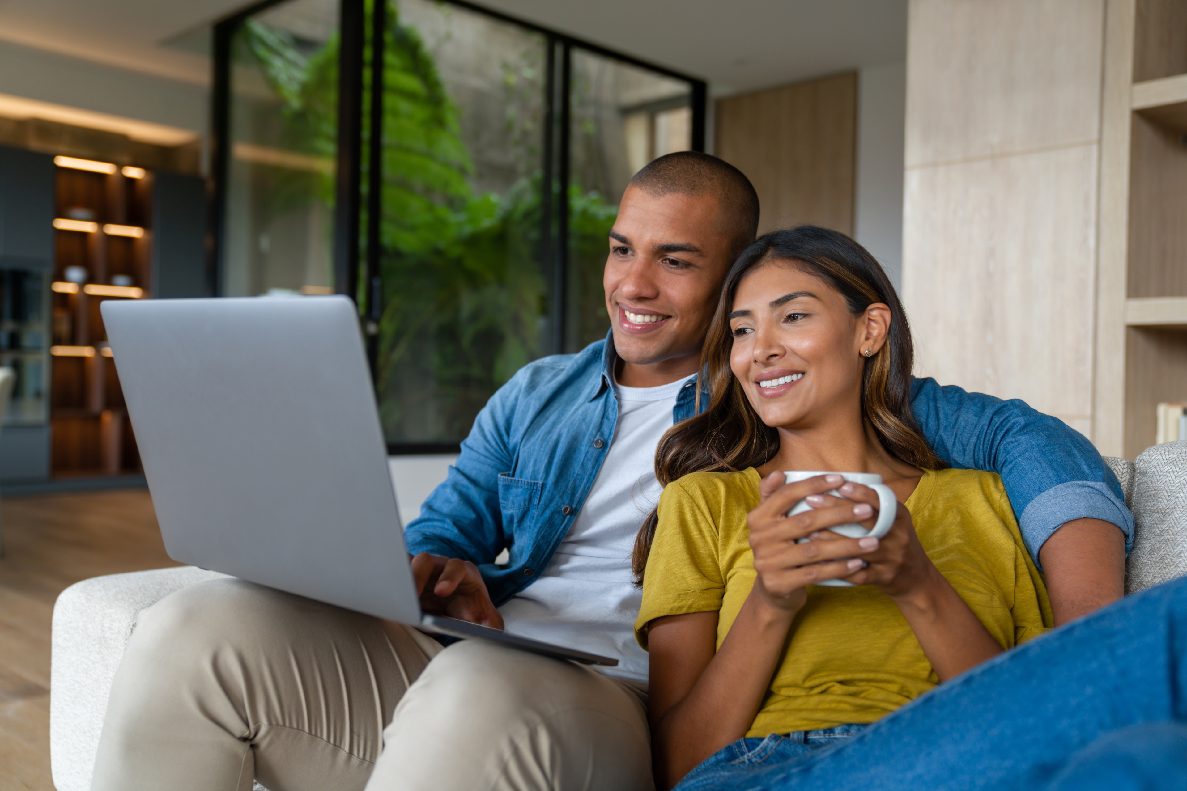 Smiling couple relaxing on a couch using a laptop together while woman holds a mug