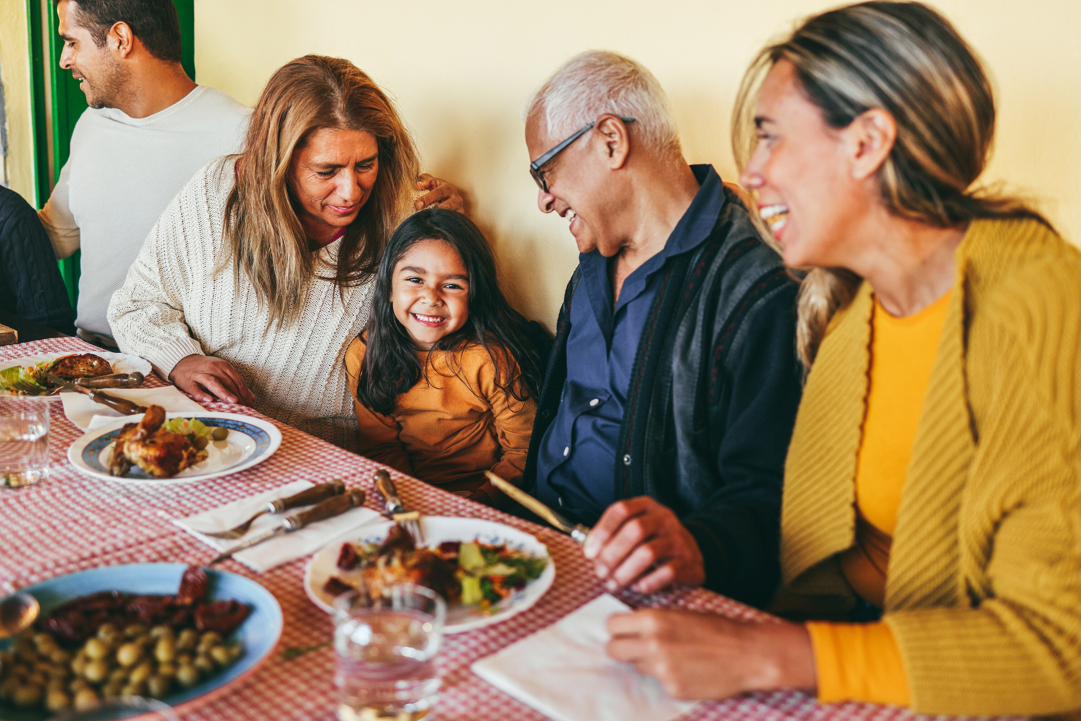 Happy multi-generational family sitting at a dining table with food, smiling together while a young girl looks up happily
