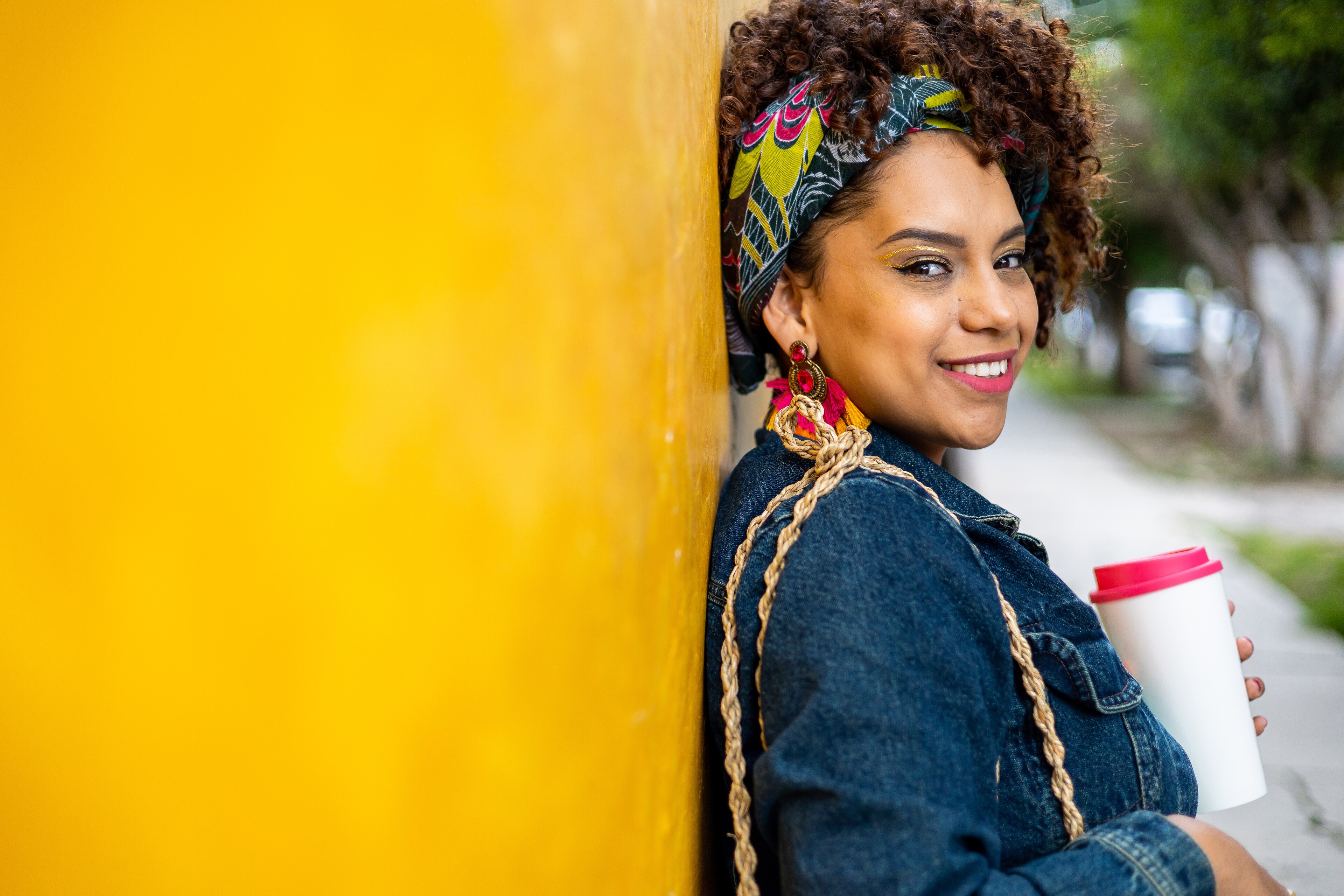 Woman with colorful headband leaning against a yellow wall holding a coffee cup