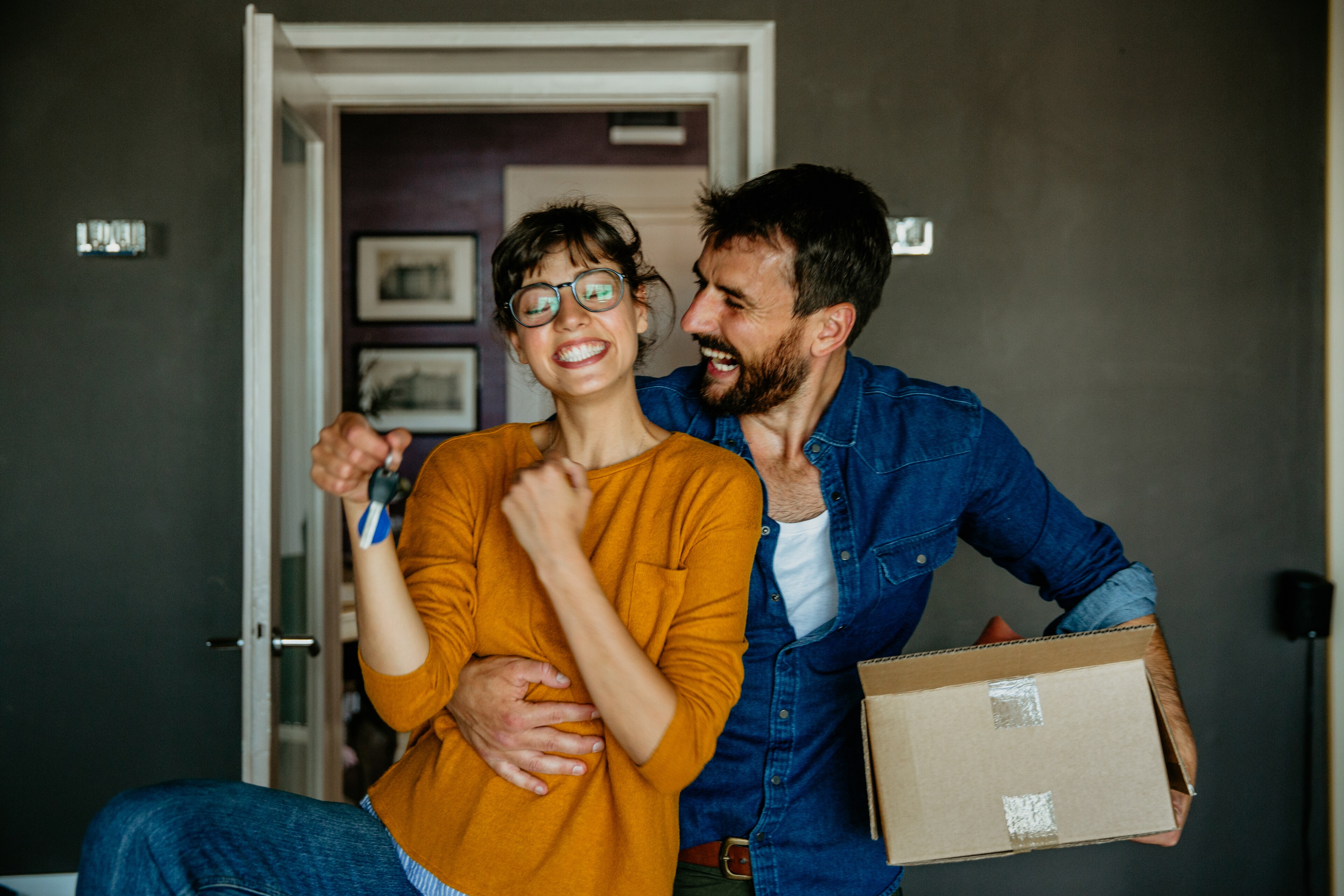 Happy young couple celebrating moving into a new home as the woman holds up house keys while the man hugs her holding an open packing box