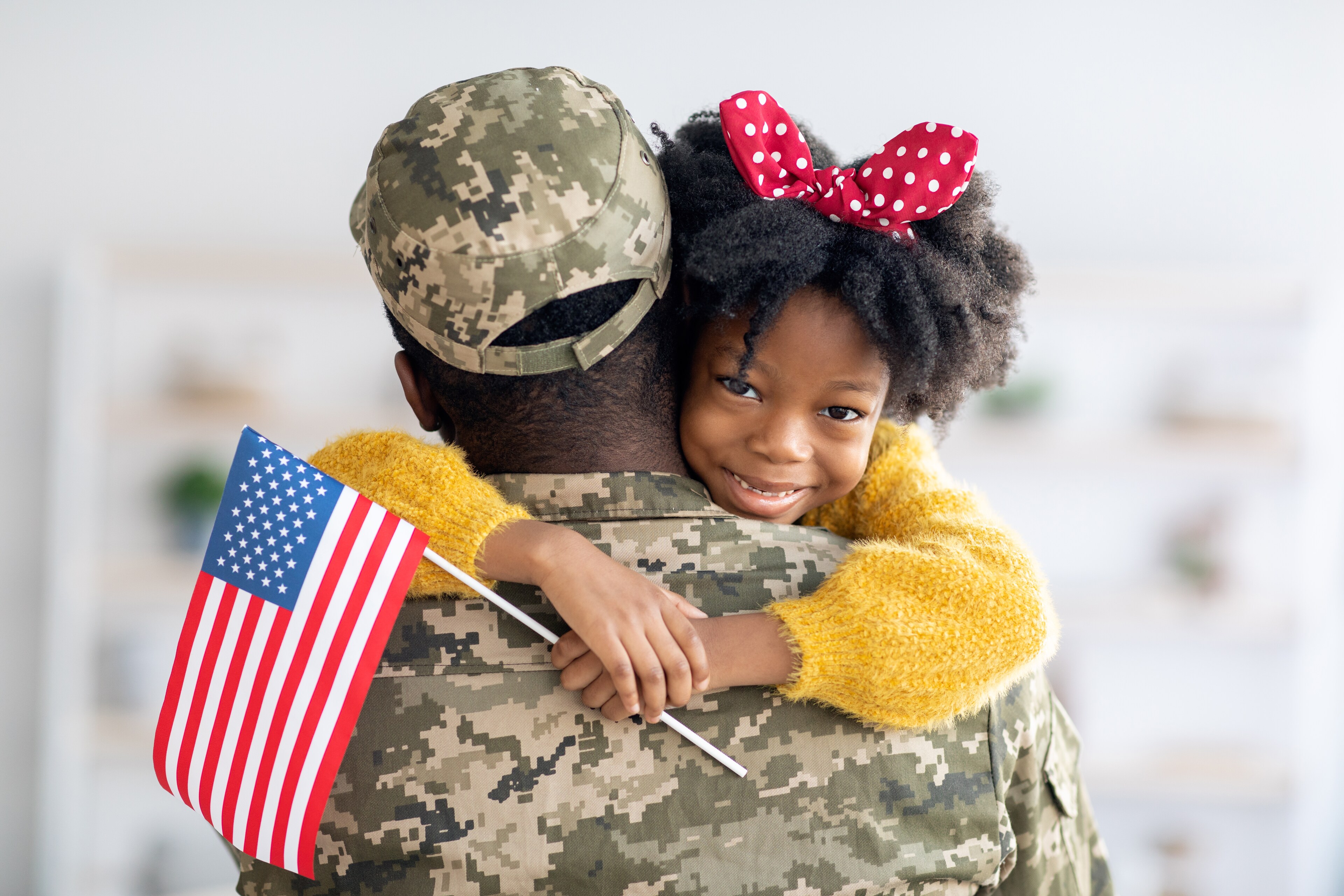 Soldier in camouflage uniform hugging a young girl holding an American flag