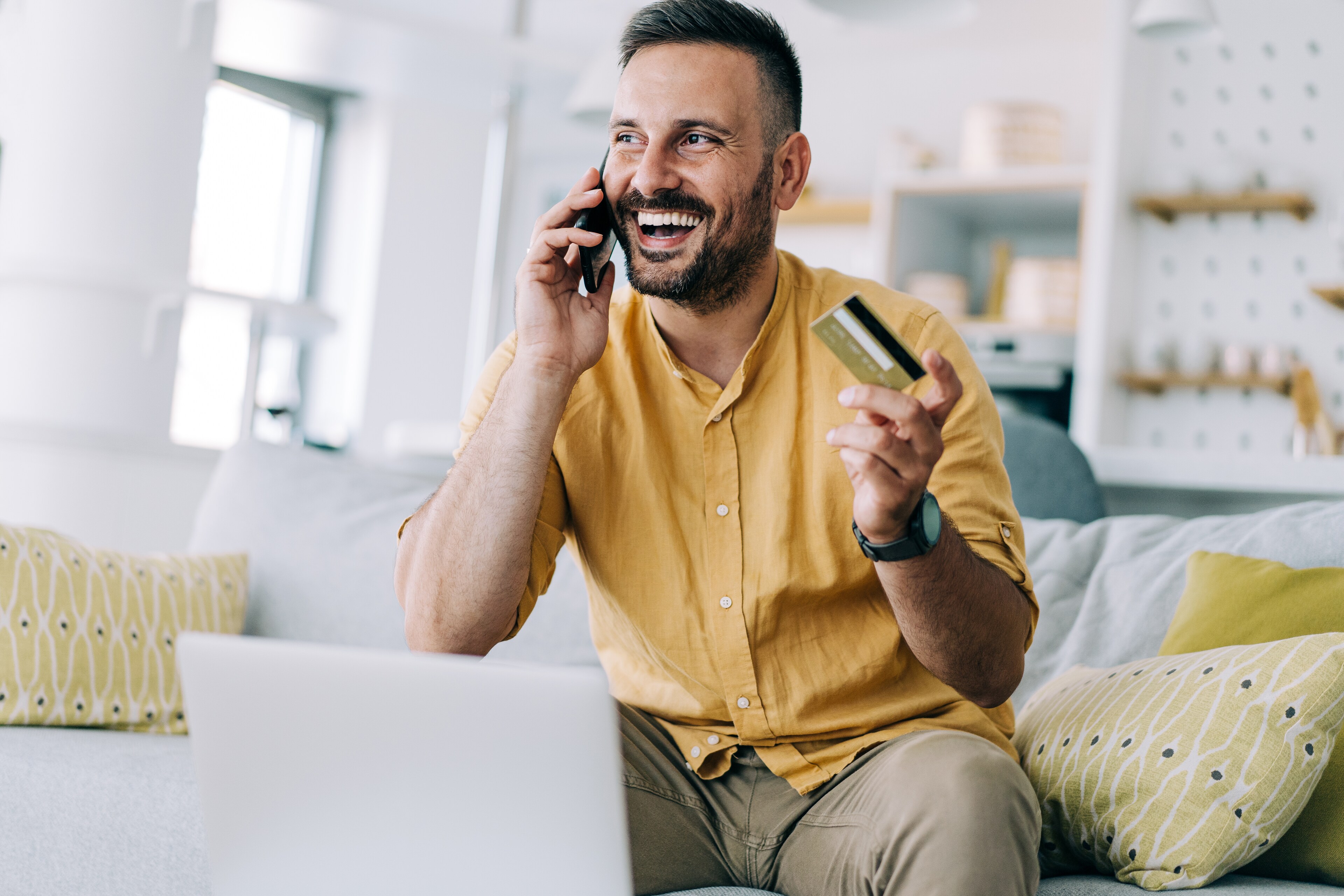 Smiling man in a yellow shirt sitting on a sofa with a laptop, holding a payment card while talking on his mobile phone