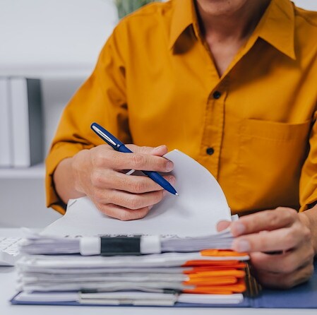 A focused office worker in a mustard shirt writes on a document while reviewing a thick stack of papers, folders, and pens at a desk.
