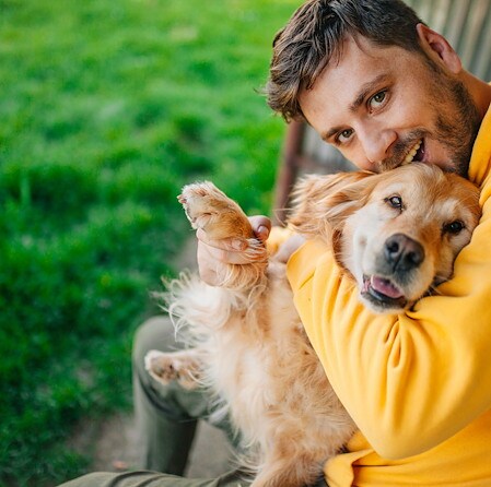Smiling man in a yellow sweater hugs a happy golden retriever outdoors on green grass; the playful dog faces the camera warmly.