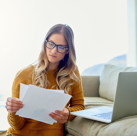 Focused young woman in glasses reads papers beside an open laptop on a sofa at home, creating a calm, studious atmosphere in warm sunlight.