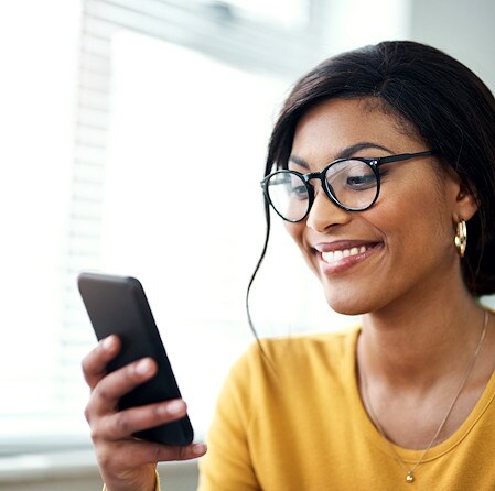 Smiling Black woman wearing glasses and a mustard-yellow top looks at her smartphone by a bright window, conveying warmth and connection.