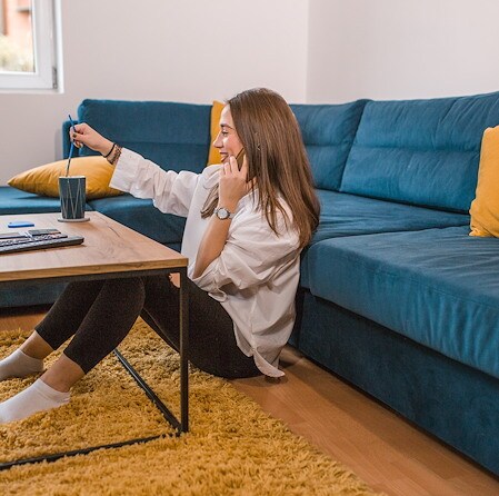 Woman relaxing on the floor beside a coffee table, holding a drink near a teal sofa with mustard pillows in a cozy living room.