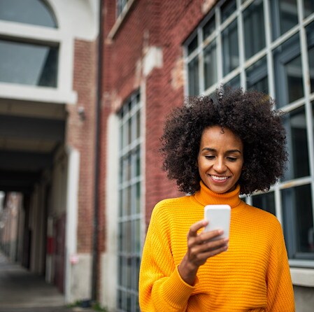 Smiling Black woman in a bright yellow sweater looks at her smartphone outside a red-brick building, conveying warmth and connection.
