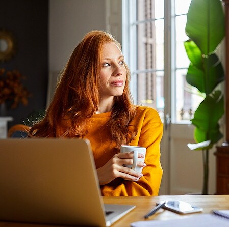 Woman sitting at a desk with a laptop holding a mug and looking out the window