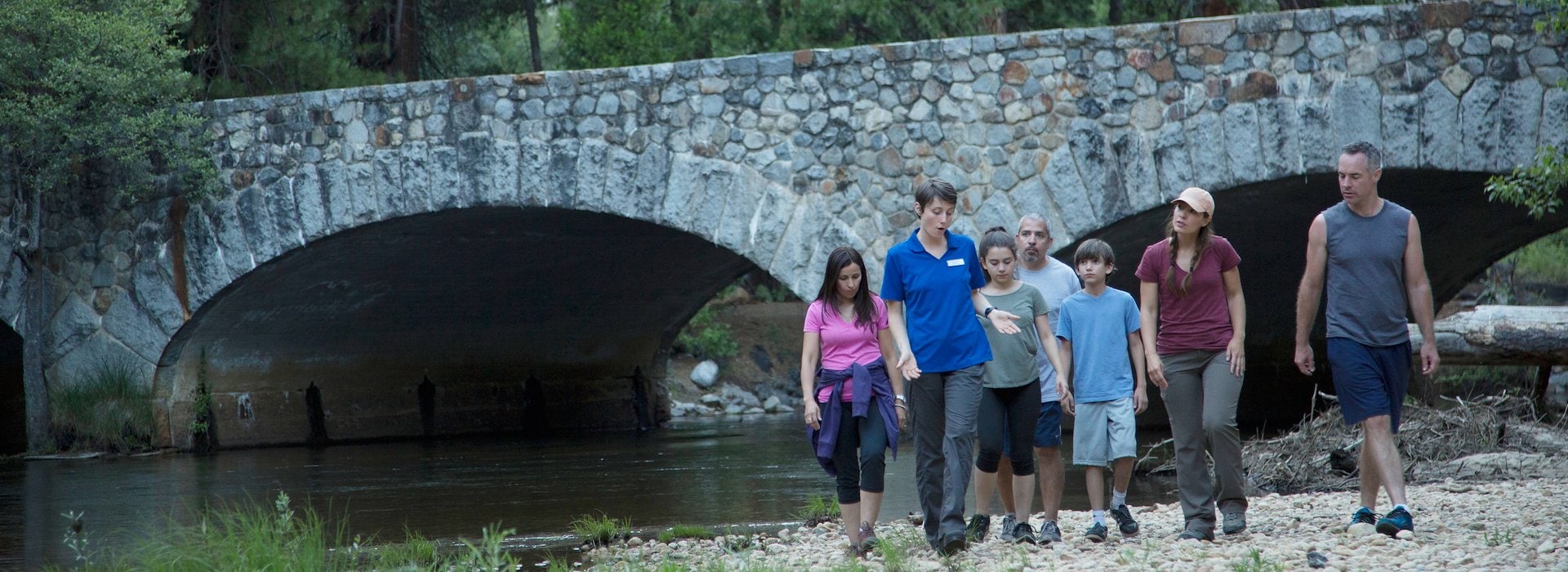 A group of people walks along a rocky path near a stone bridge over a serene river Yosemite National Park Lodging and Activities - California - United States