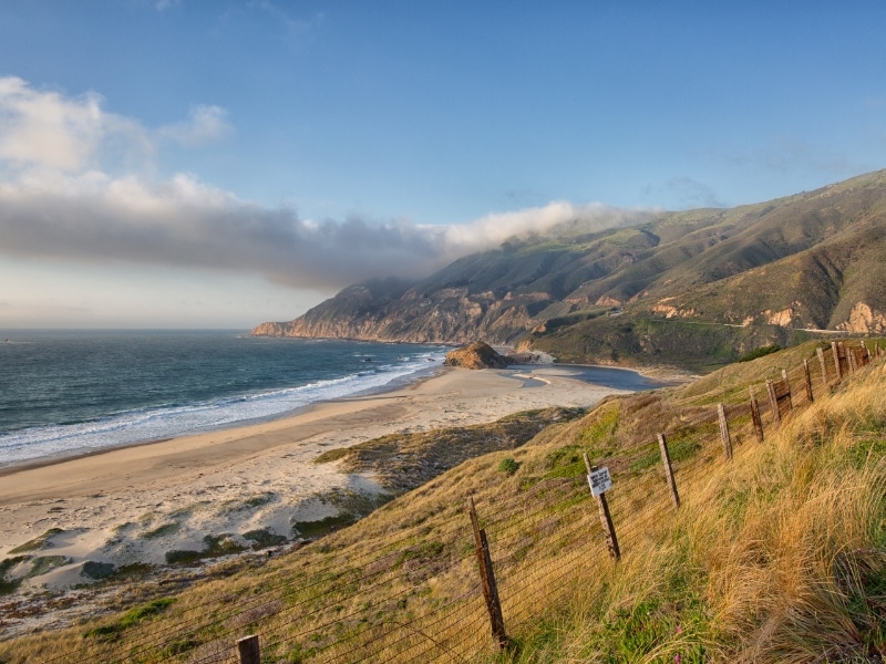 A scenic view of a beach with a sandy shore, mountains in the background, and a cloudy sky.