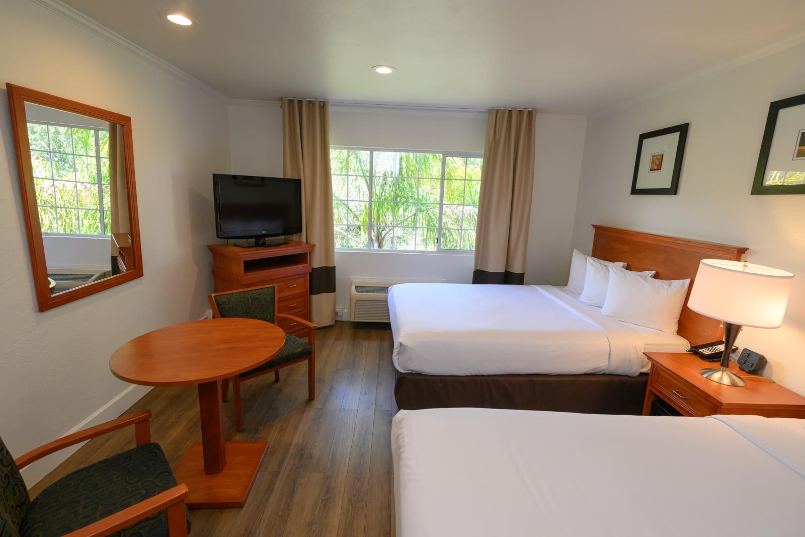 A hotel room with two beds, a wooden table and chairs, a TV on a stand, and a window with curtains.
