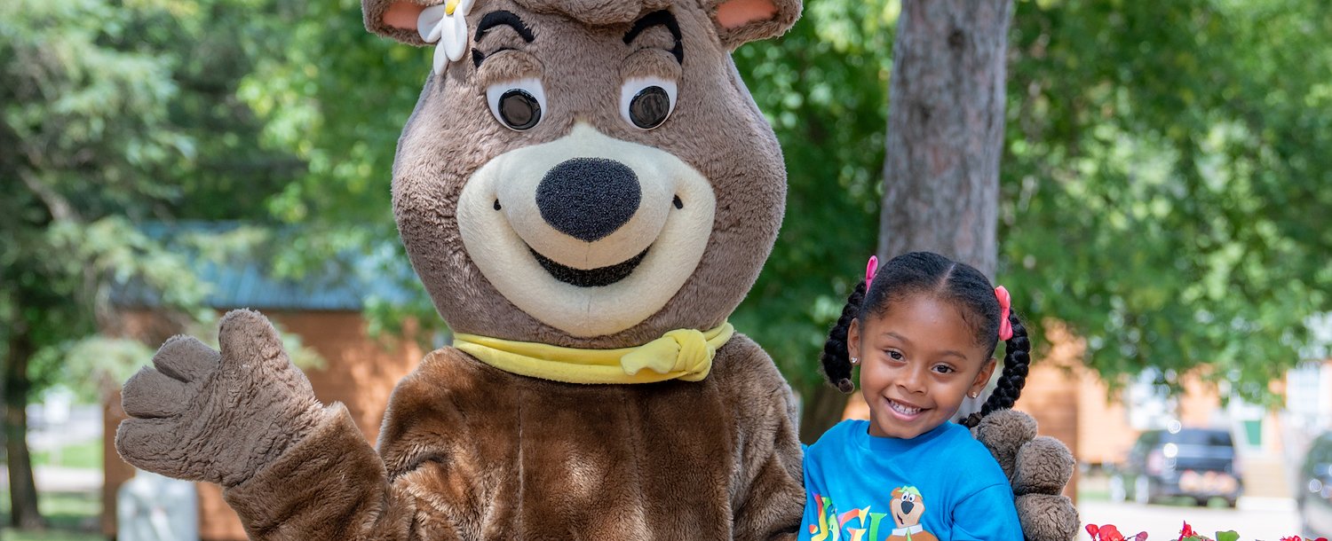 A girl sitting on a bench with Yogi Bear at Jellystone Park™ Williamsburg