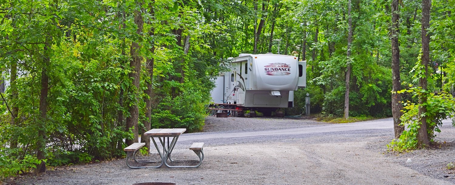Campsite with a picnic table, fire ring, and an RV parked among trees at Jellystone Park™ Williamsburg
