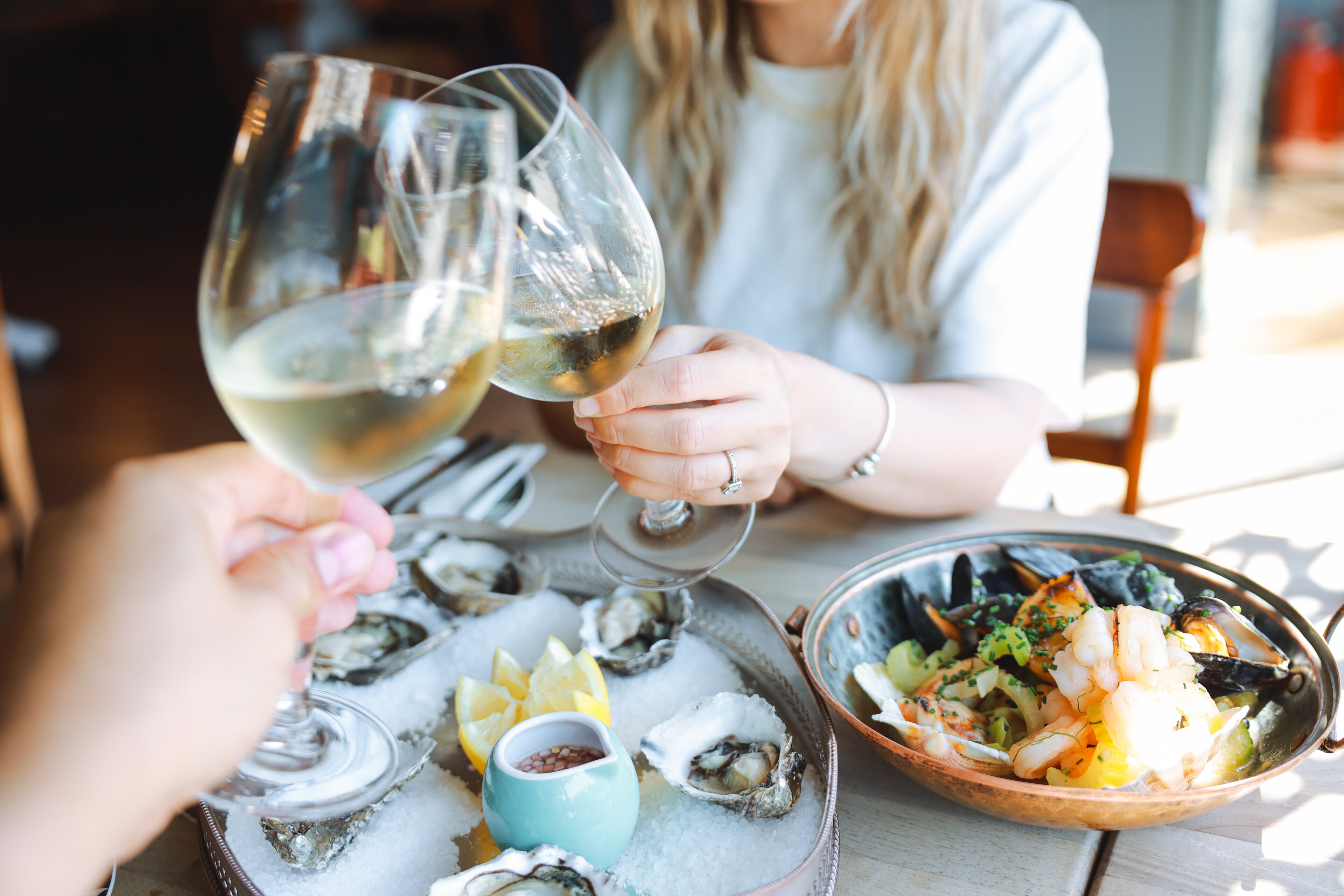 Two people toast with white wine glasses over a seafood spread featuring oysters on ice and a cooked shellfish dish.