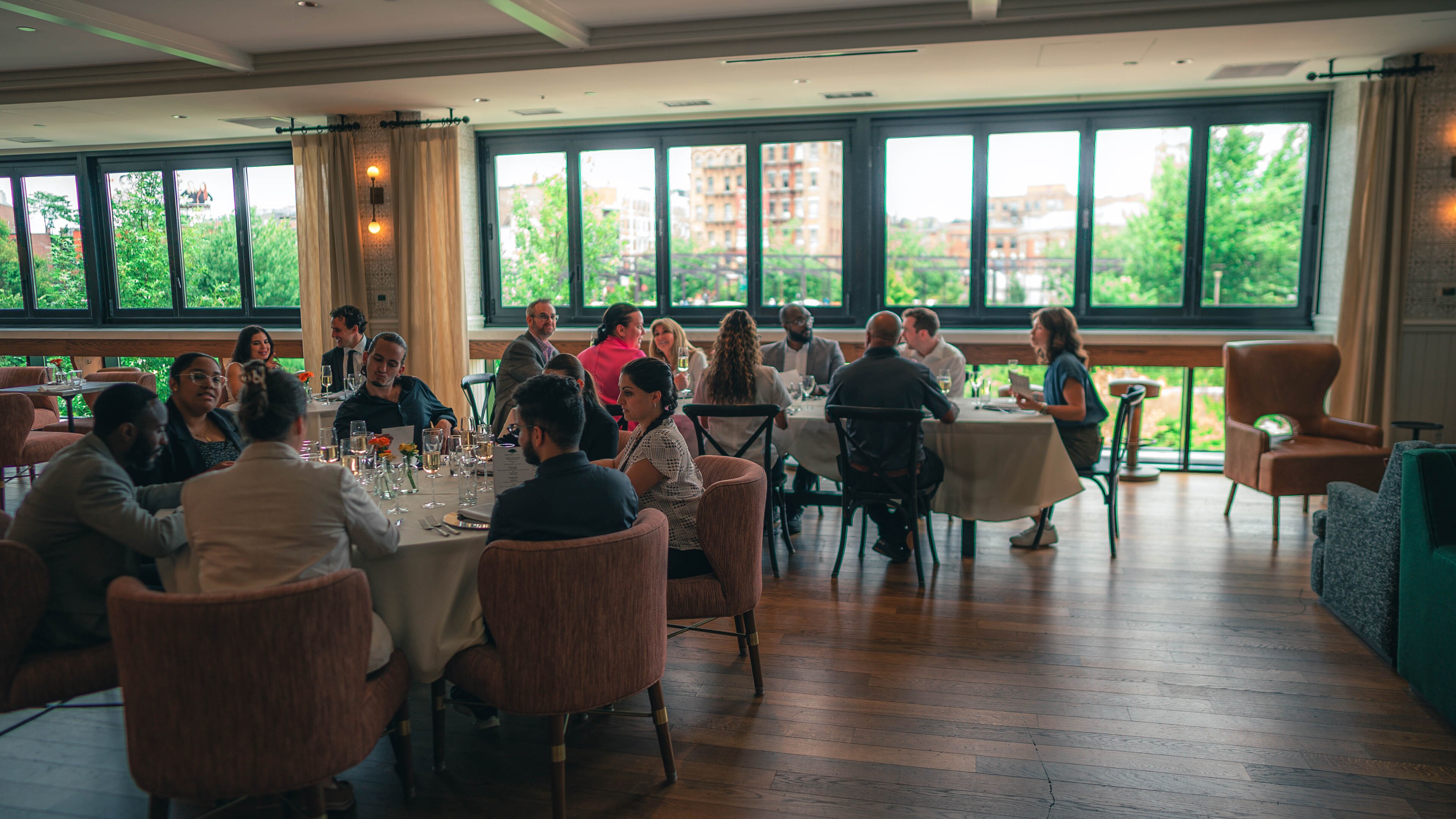 People seated around dining tables, enjoying conversation during a social gathering in a bright event space with large windows overlooking greenery.
