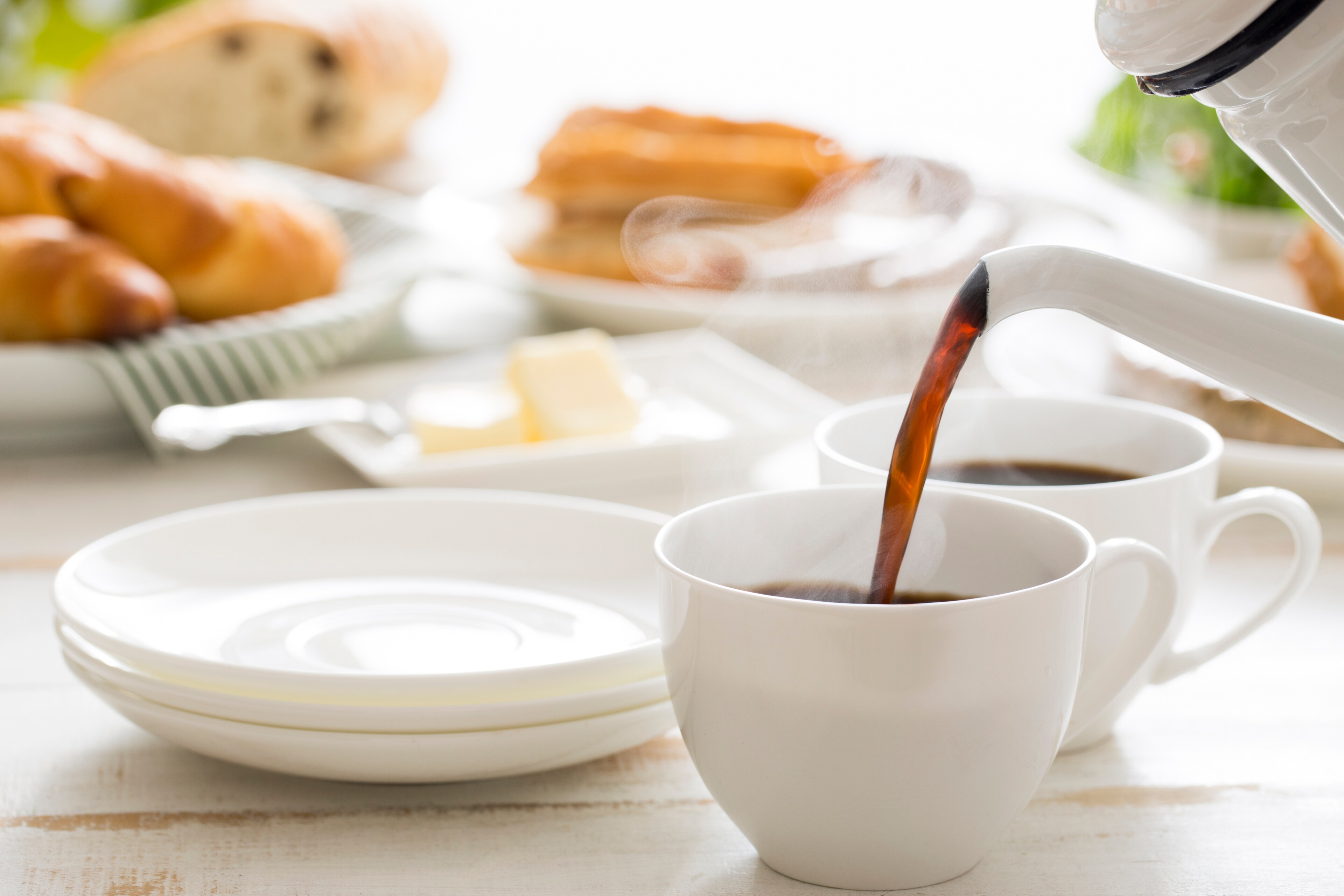 Hot coffee being poured into a white cup with breakfast pastries and bread in the background