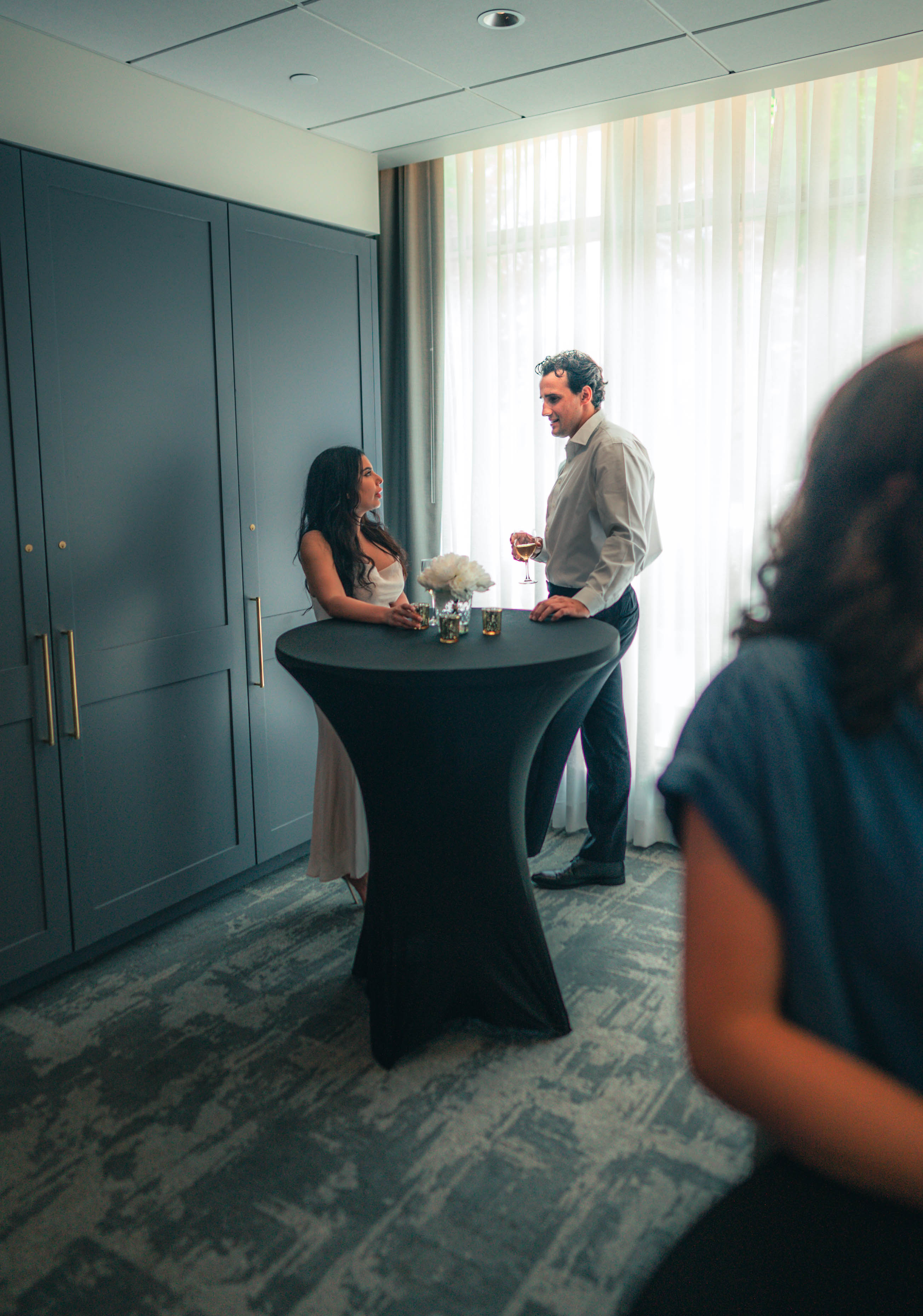 A man and woman stand by a cocktail table, chatting with drinks in hand near a large window at an indoor gathering.