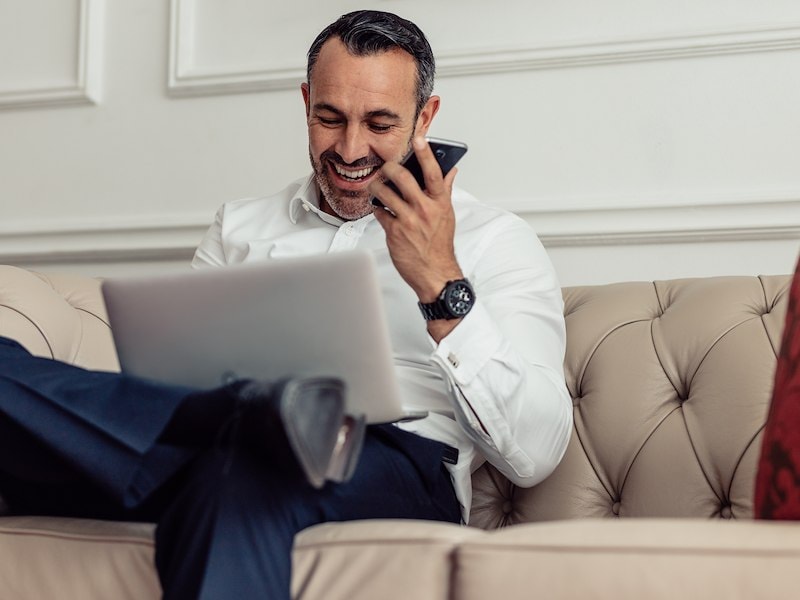 A man sitting on a sofa in a lobby talking on the phone and using a laptop