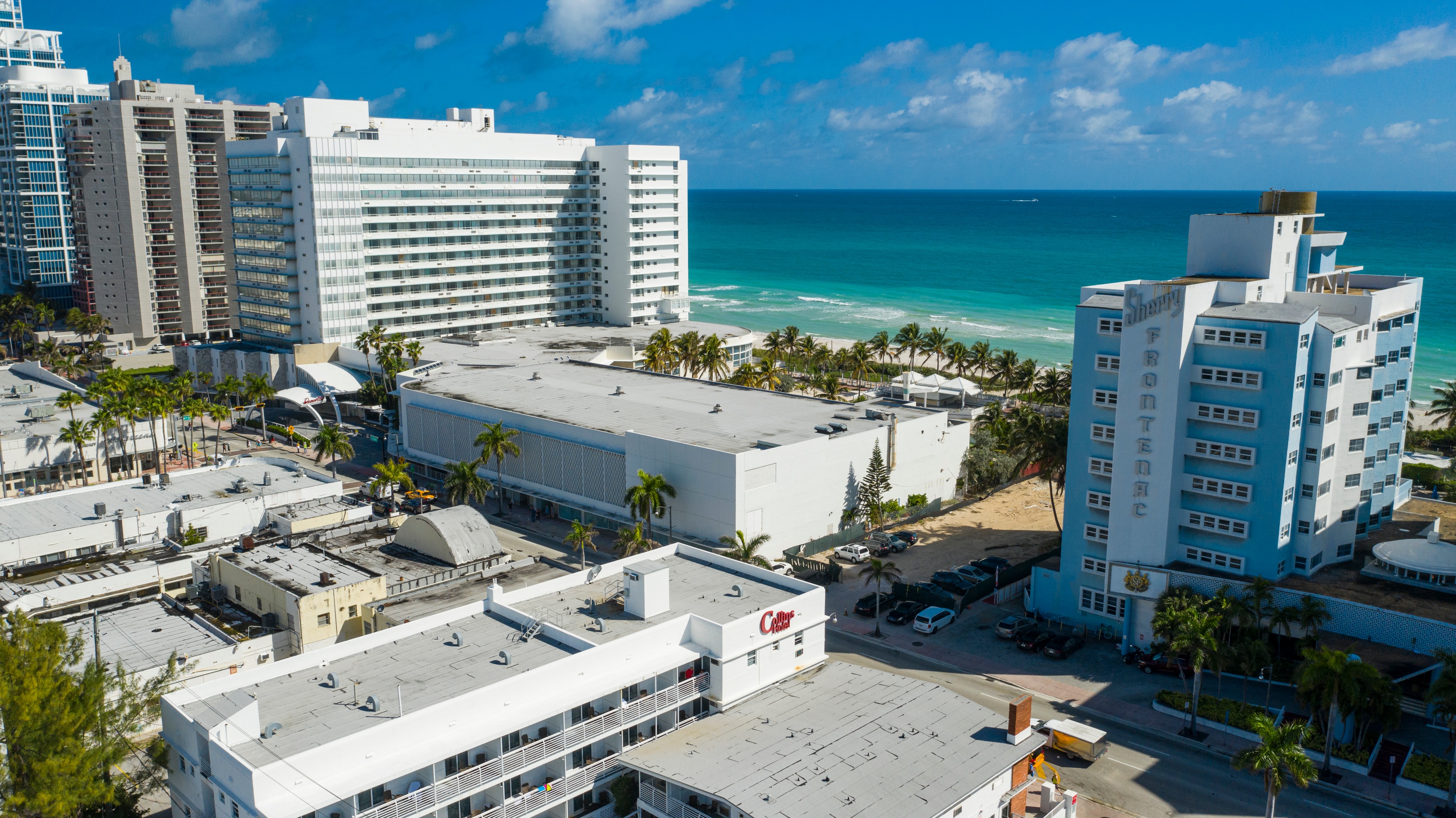 An aerial view of a beachfront area with several white buildings, a parking lot, and a large body of water in the background.