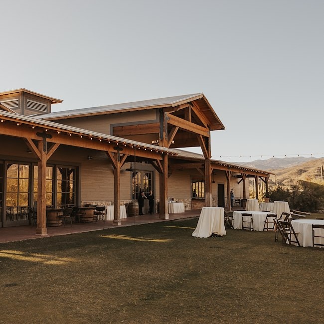 A large building with a wooden exterior and a large overhang sits on a grass lawn with tables and chairs set up outside.