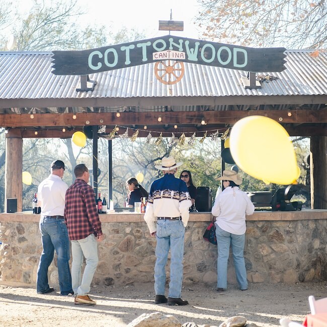 A rustic outdoor bar with a corrugated metal roof and stone counter, surrounded by people.