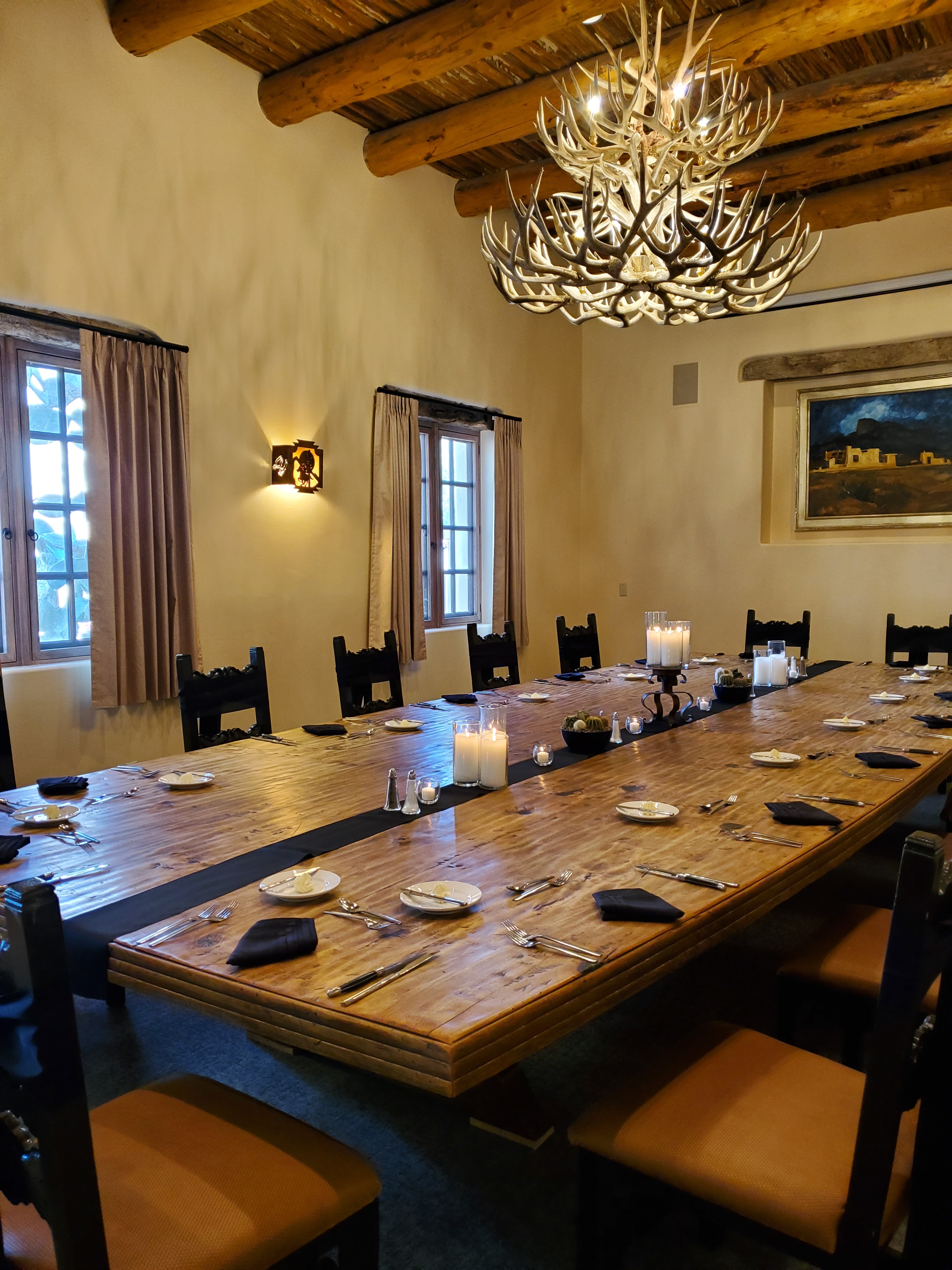 A formal dining room with a long wooden table set for a large group, adorned with an antler chandelier and candlelight.