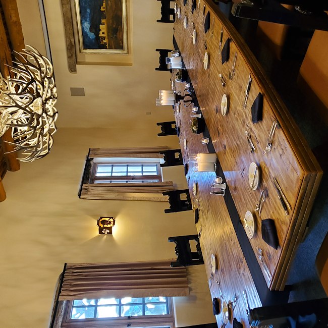 A formal dining room with a long wooden table set for a large group, adorned with an antler chandelier and candlelight.