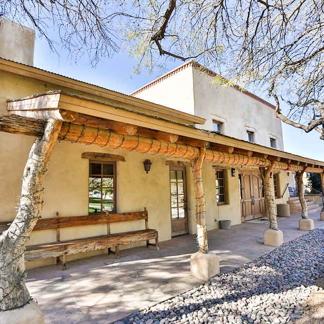 A southwestern-style building with stucco walls, wooden beams, and a clay tile roof, featuring a large arched porch with benches.