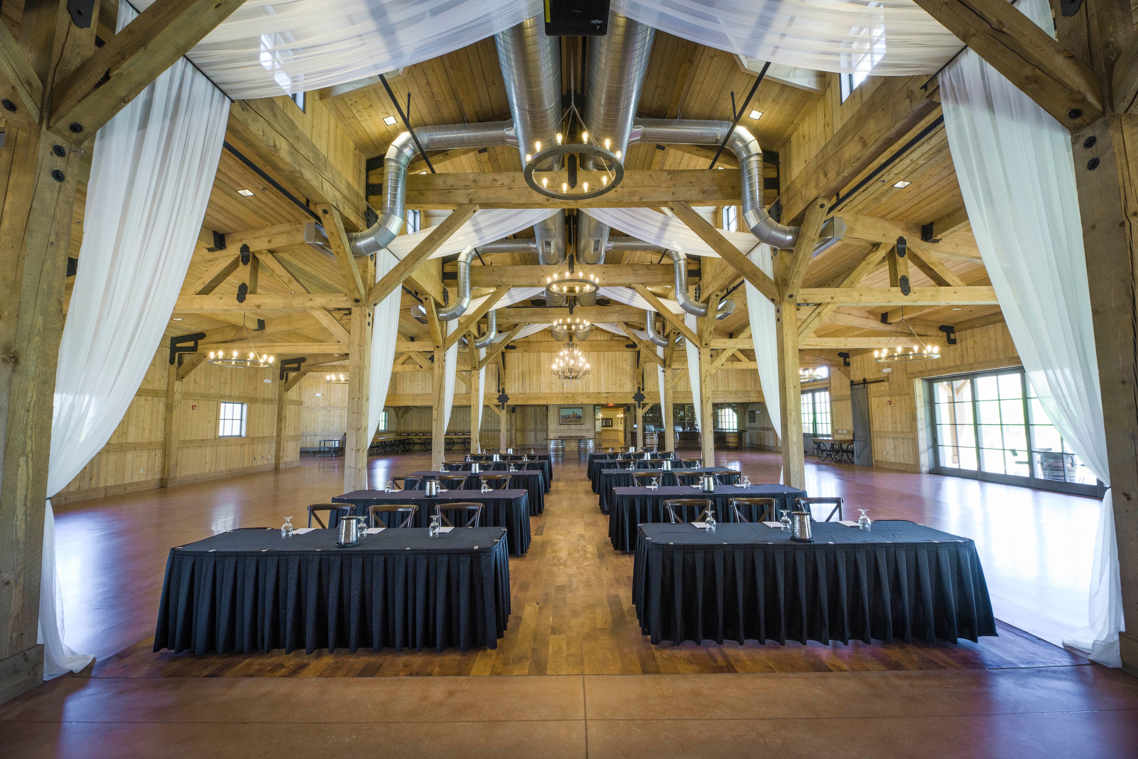 A large, open room with wooden beams, white curtains, and round tables with black tablecloths.