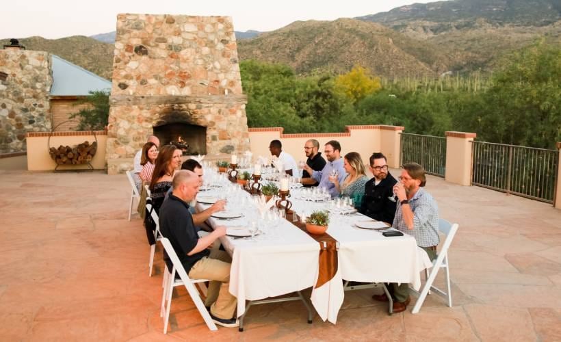 A group of people sitting around a long table with white tablecloths, set with plates, glasses, and silverware, in front of a stone fireplace with a mountain view.