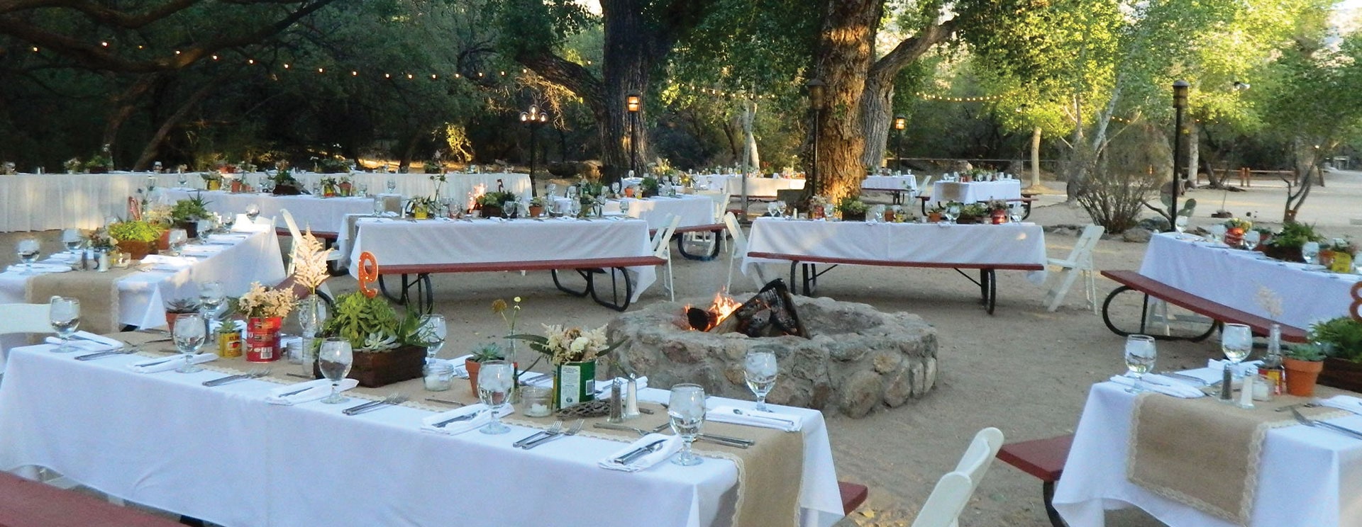 An outdoor dining area with tables set for a meal, a fire pit, and trees in the background.